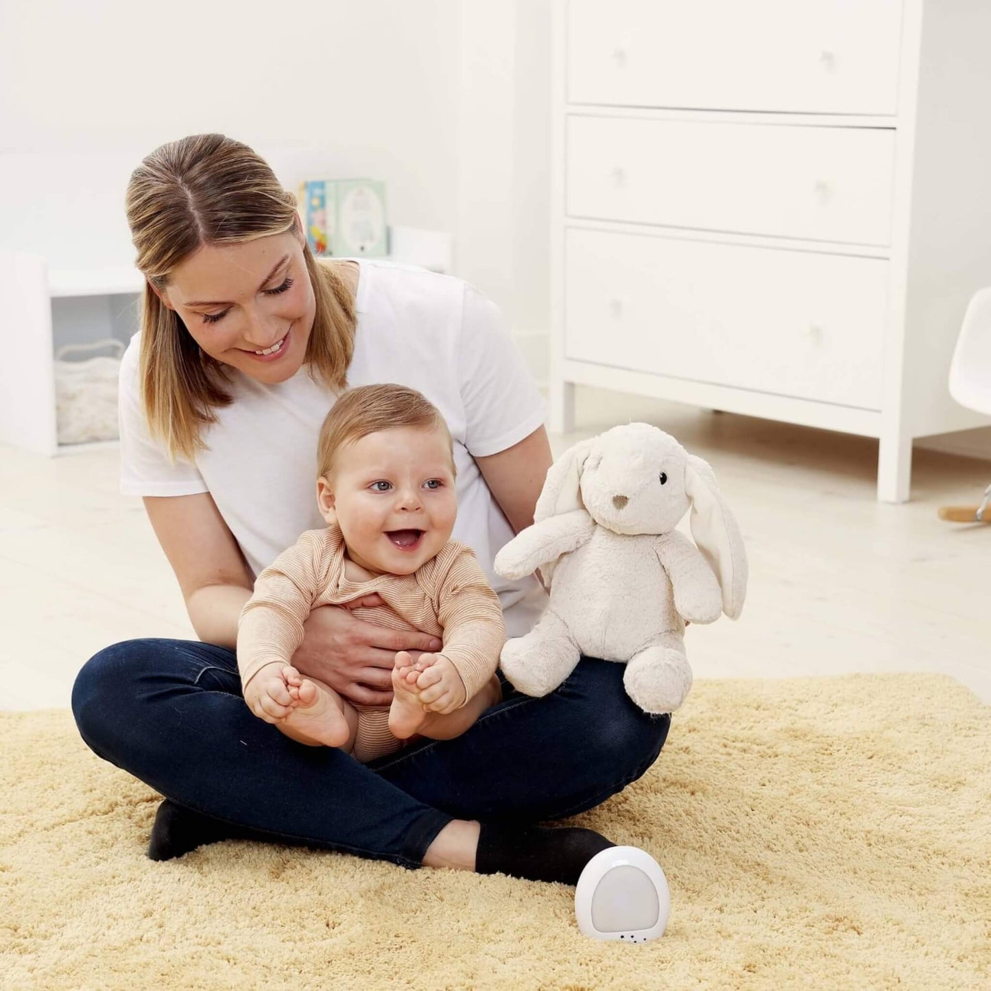 Mother sits on the floor with a smiling baby on her lap, while a plush bunny toy rests beside them and a night-light unit sits on the rug.