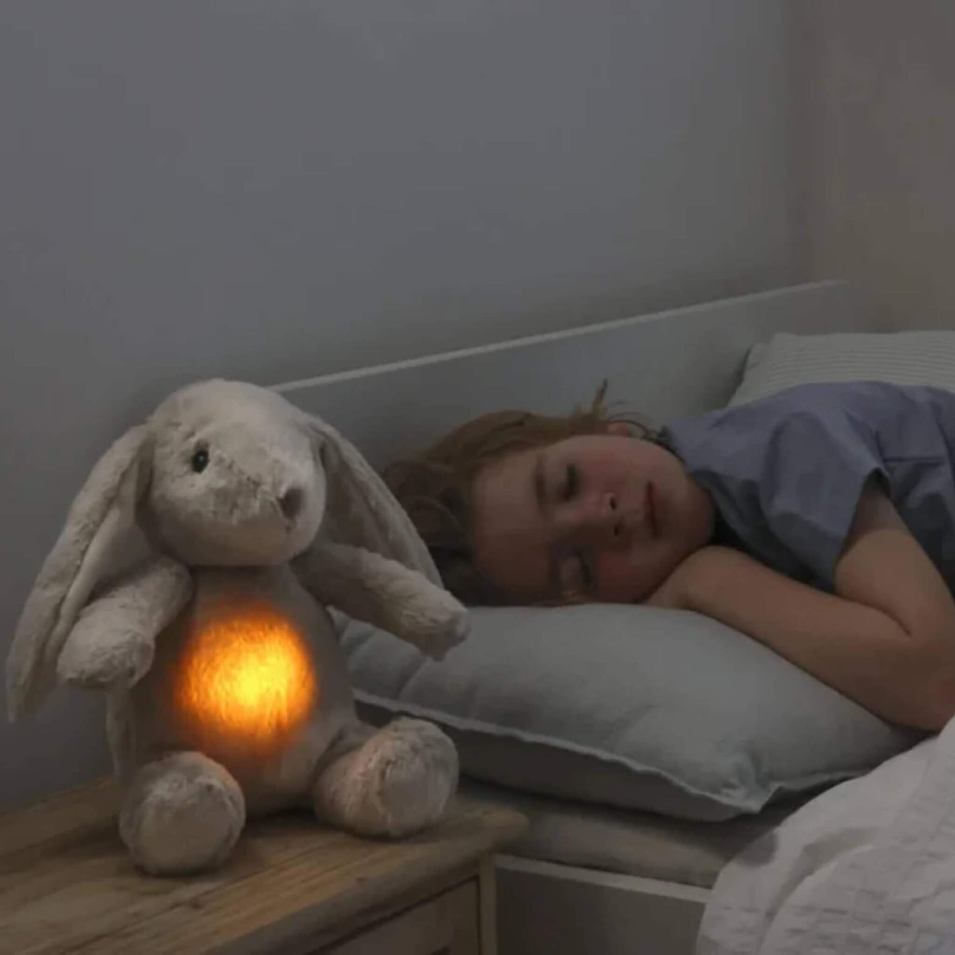Child in a blue top sleeps in bed while a plush bunny toy with a glowing tummy sits on the bedside table.