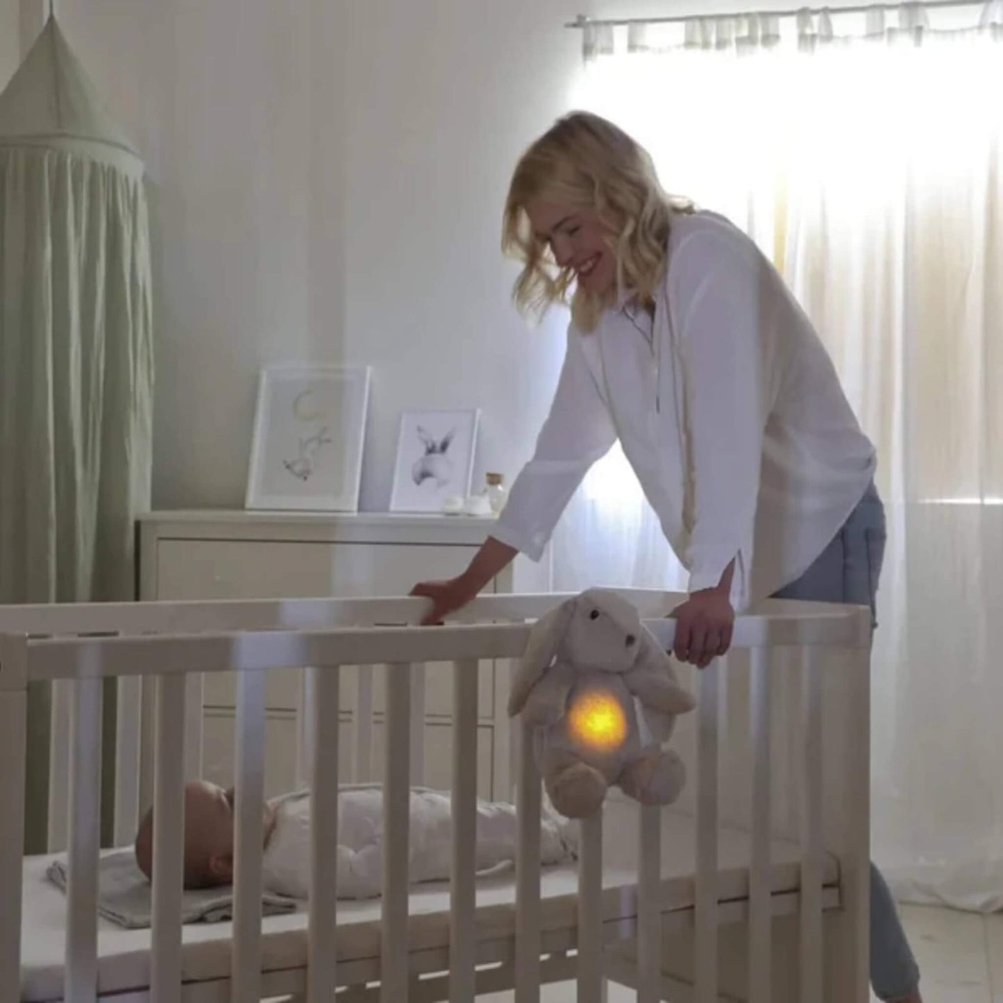 Woman leans over a cot smiling at a baby while a plush bunny toy with glowing tummy hangs from the cot rail.