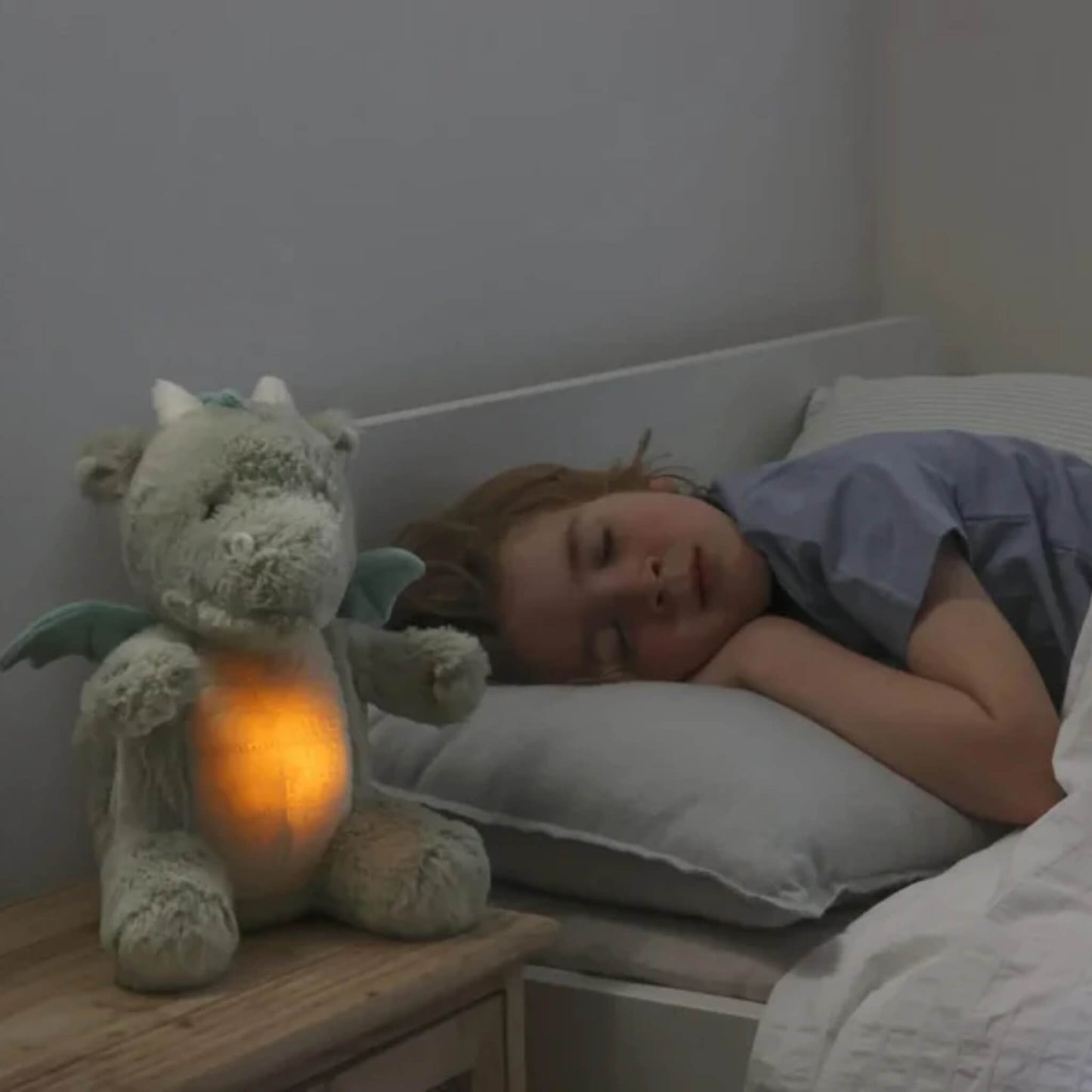 A child sleeps on a pillow while the plush dragon toy sits on a bedside table glowing orange, softly lighting the room.