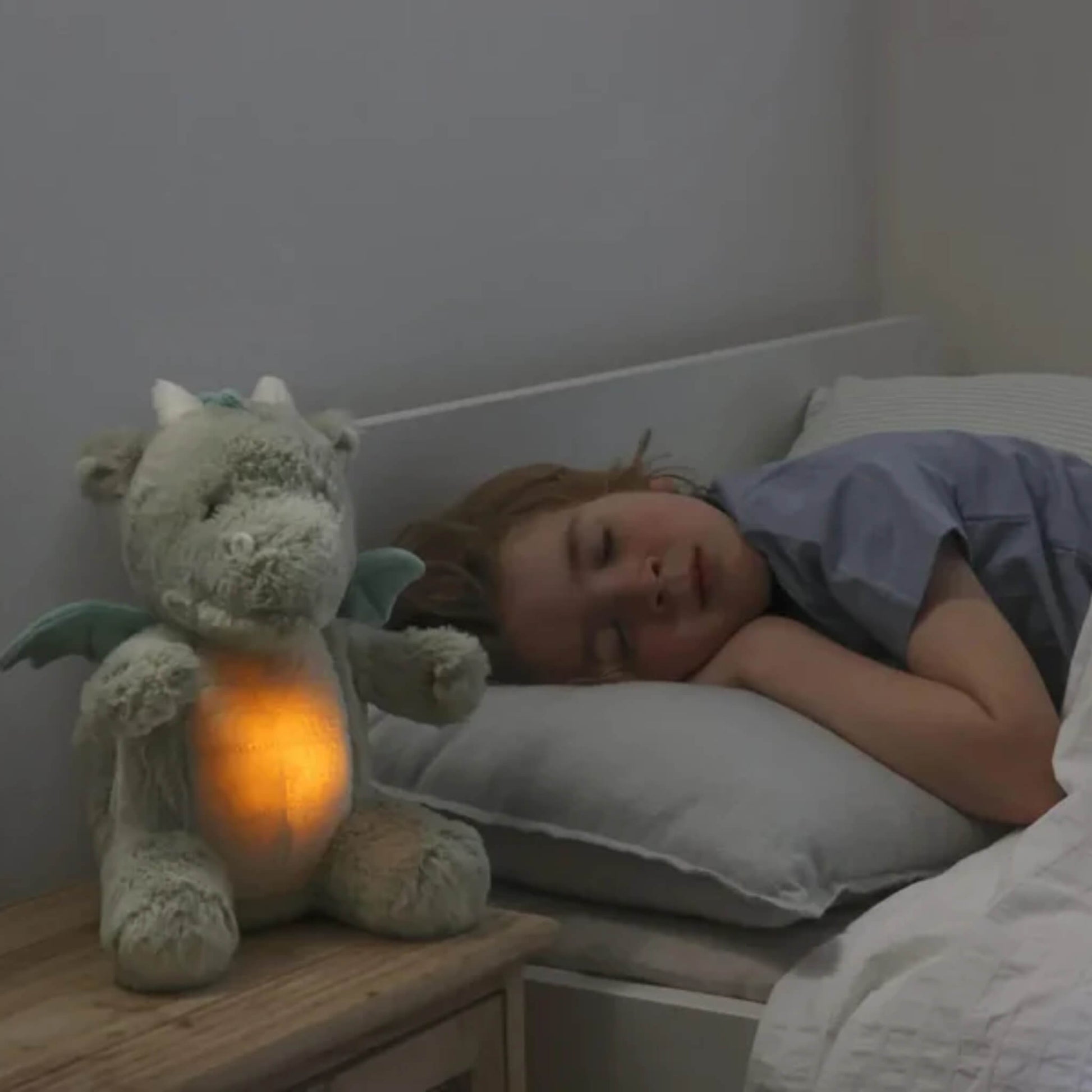 A child sleeps on a pillow while the plush dragon toy sits on a bedside table glowing orange, softly lighting the room.