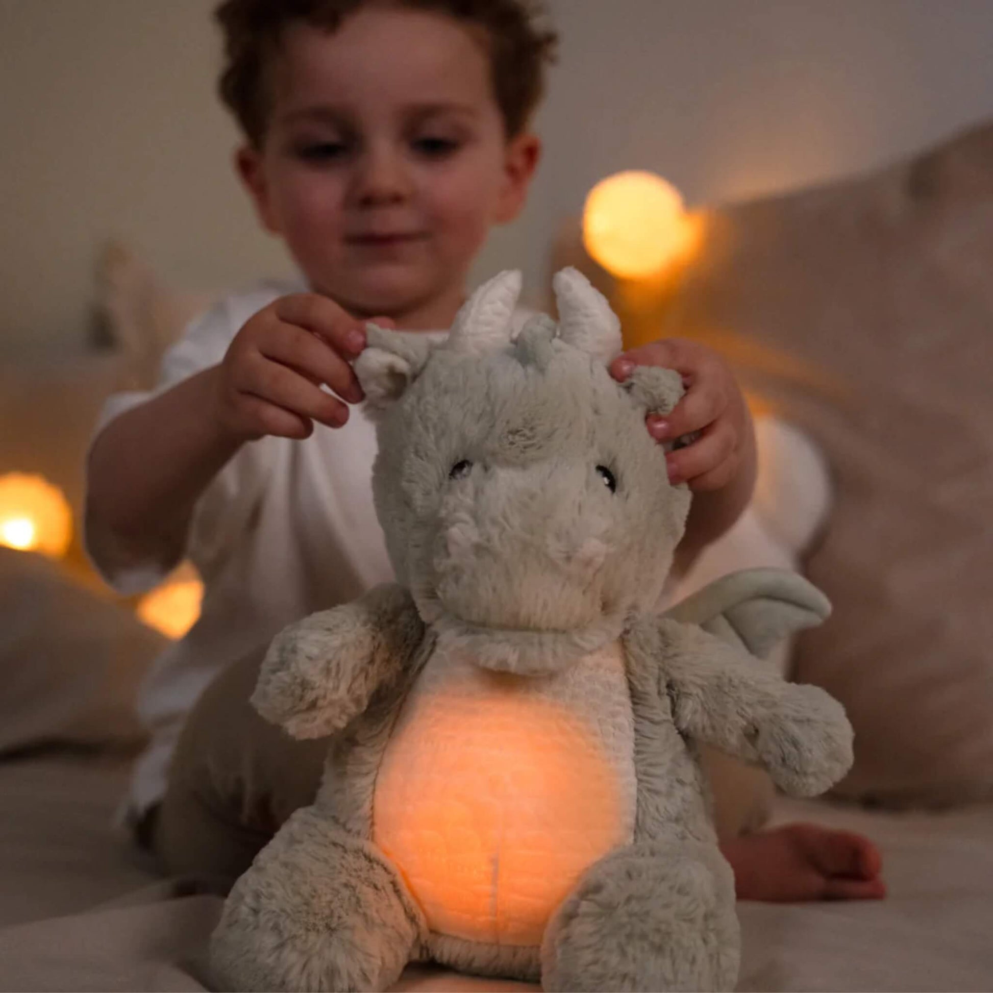 A young boy sits on a bed playing with a plush dragon toy, its tummy glowing orange while warm lights shine in the background.
