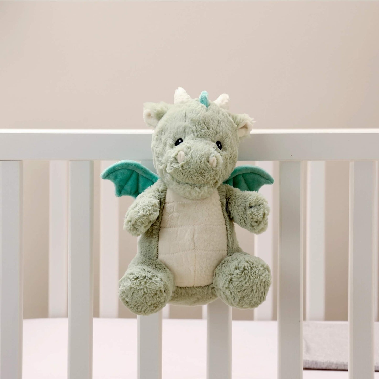 The plush dragon toy hangs securely on the side of a white baby cot using its attached strap.