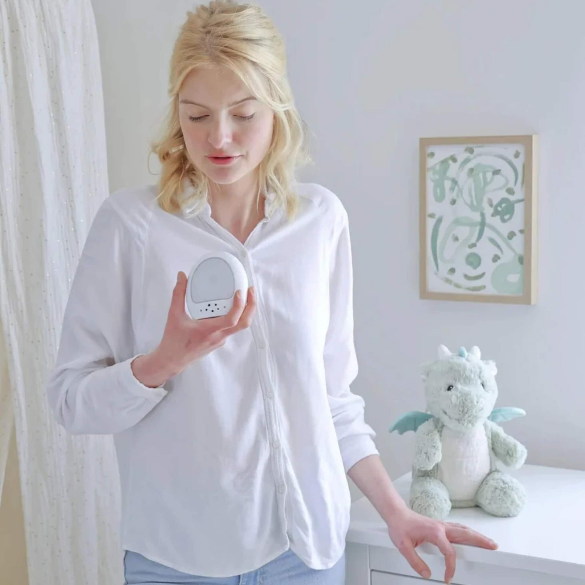 A woman in a white shirt holds the nightlight device while the plush dragon toy rests on a nursery dresser in the background.