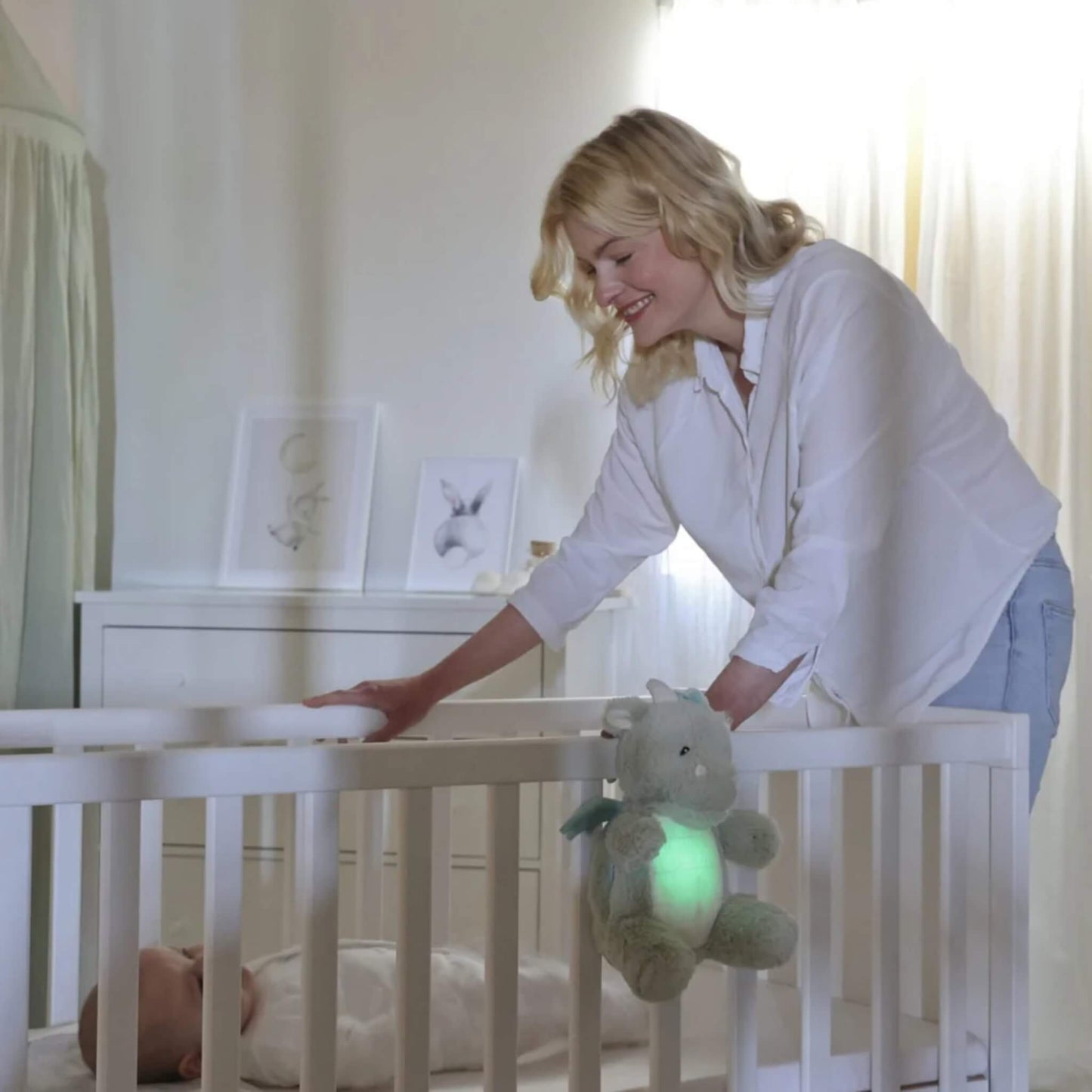 A woman smiles while standing by a cot where a baby lies, as the plush dragon toy glows green while hanging on the cot rail.