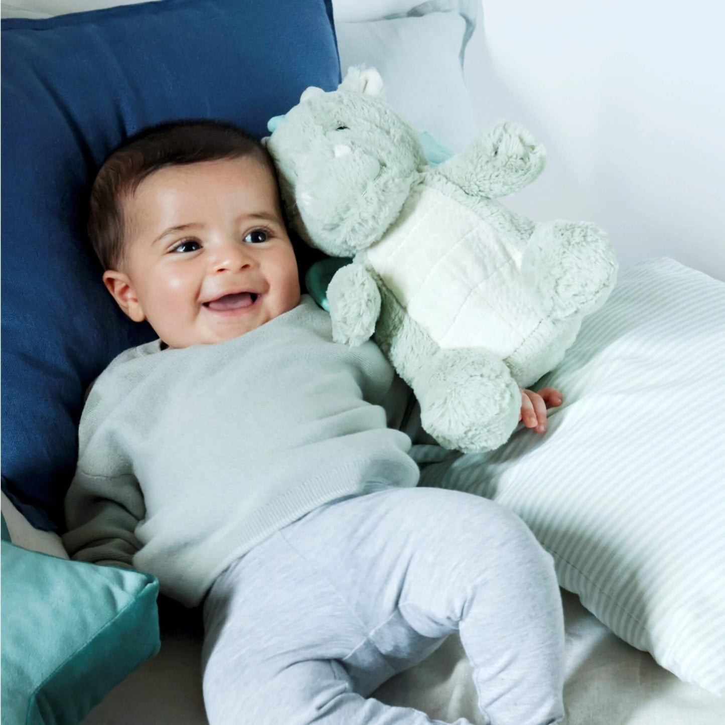 A smiling baby lies on a bed holding the plush dragon toy close, looking comfortable and happy.
