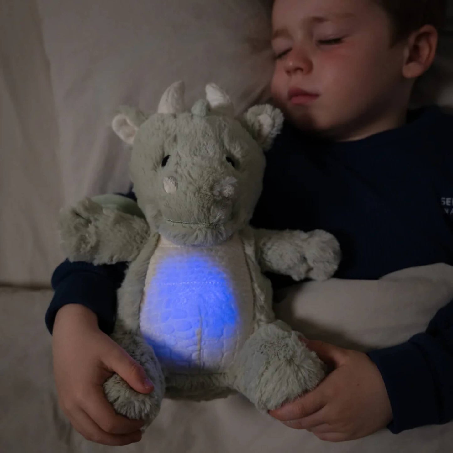 A young child sleeps in bed while holding the plush dragon toy, its tummy glowing blue in the dark room.