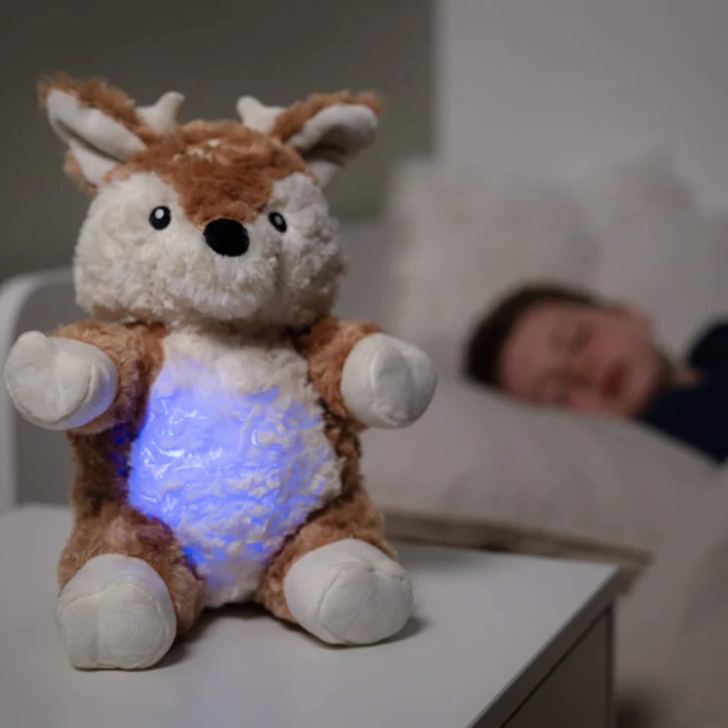 Soft fawn plush toy with glowing blue tummy placed on a bedside table while a child sleeps in the background.