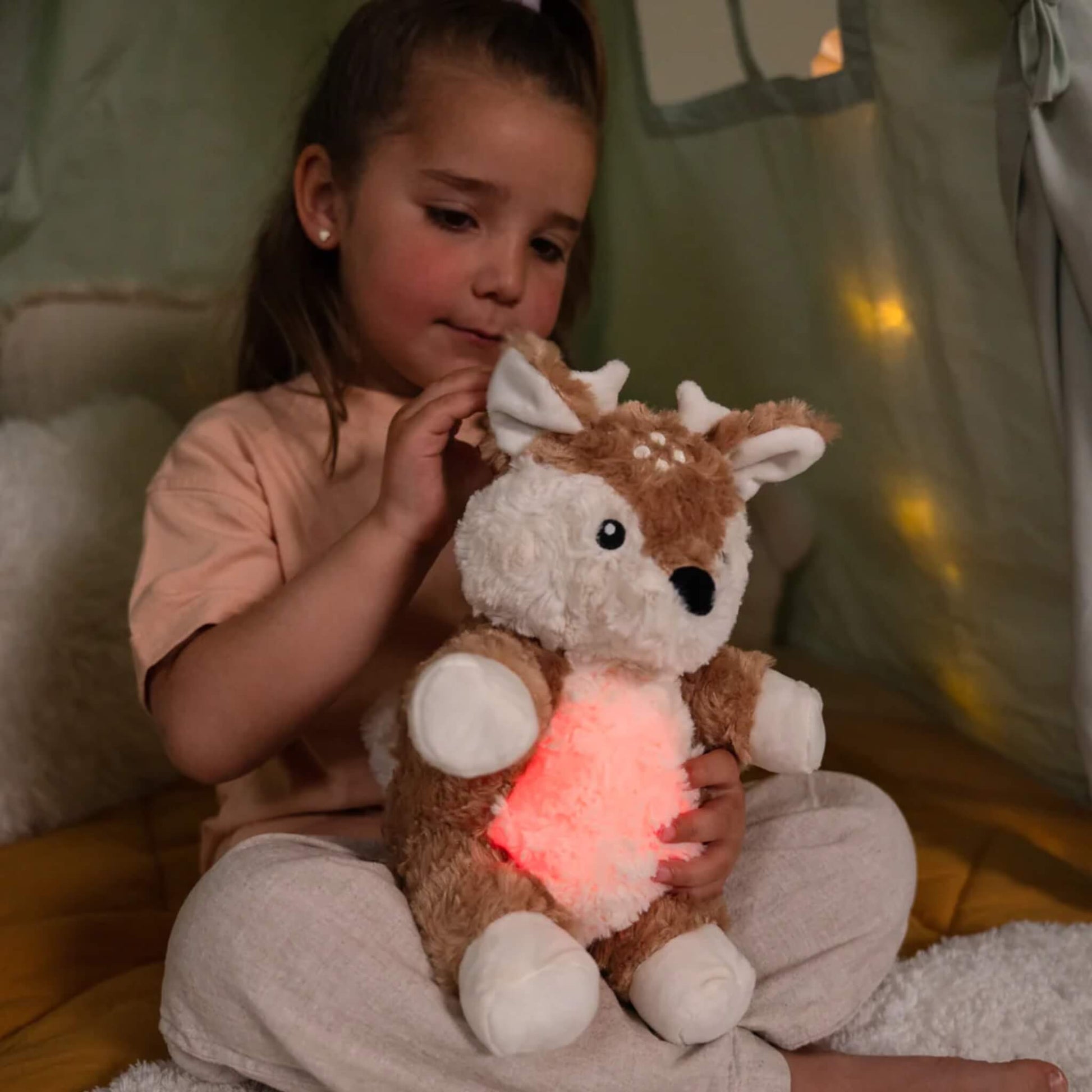 Girl sitting on a soft mat inside a play tent, holding a fawn plush toy with its tummy glowing red.