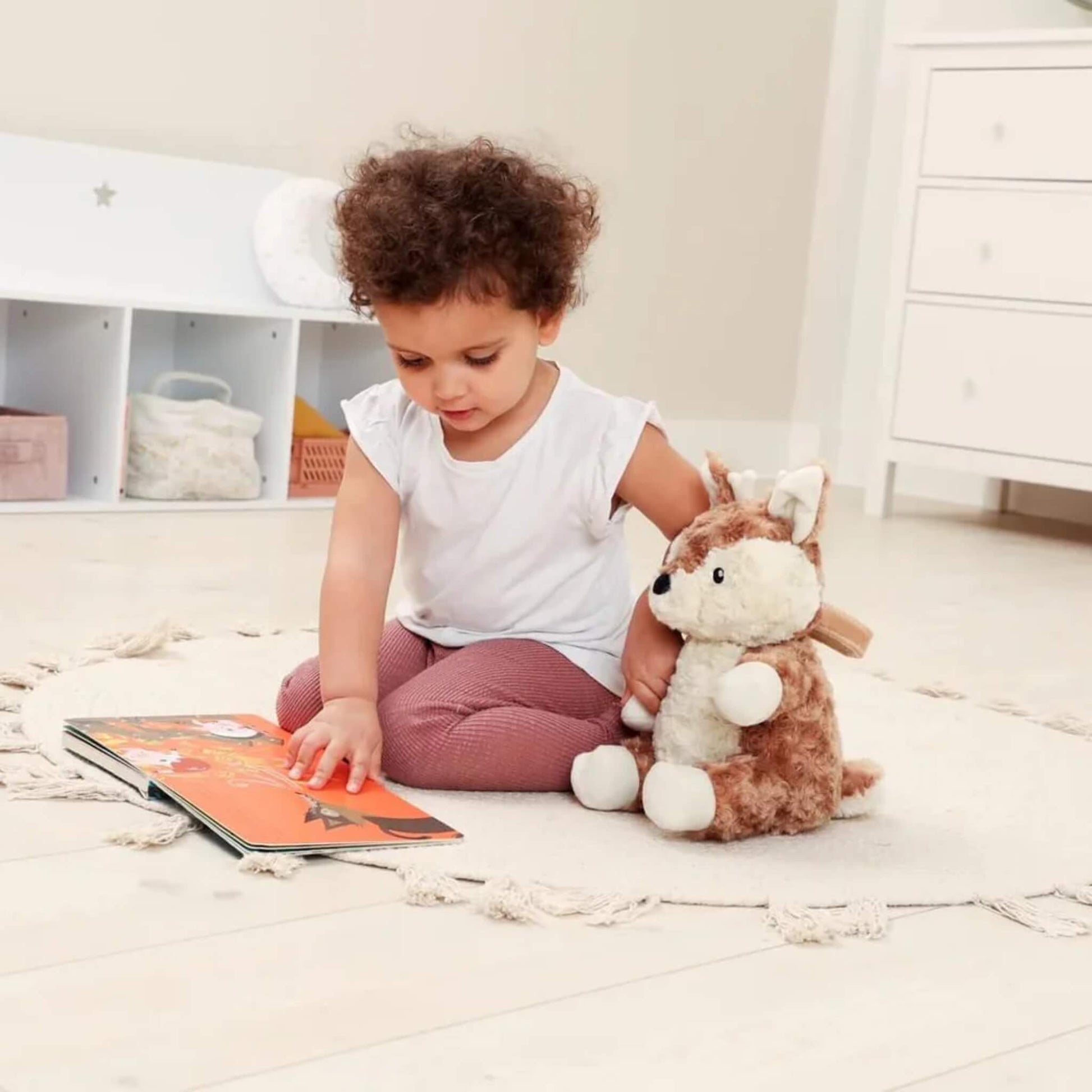 Child sitting cross-legged on a rug, pointing at a picture book while a soft fawn plush toy rests at their side.