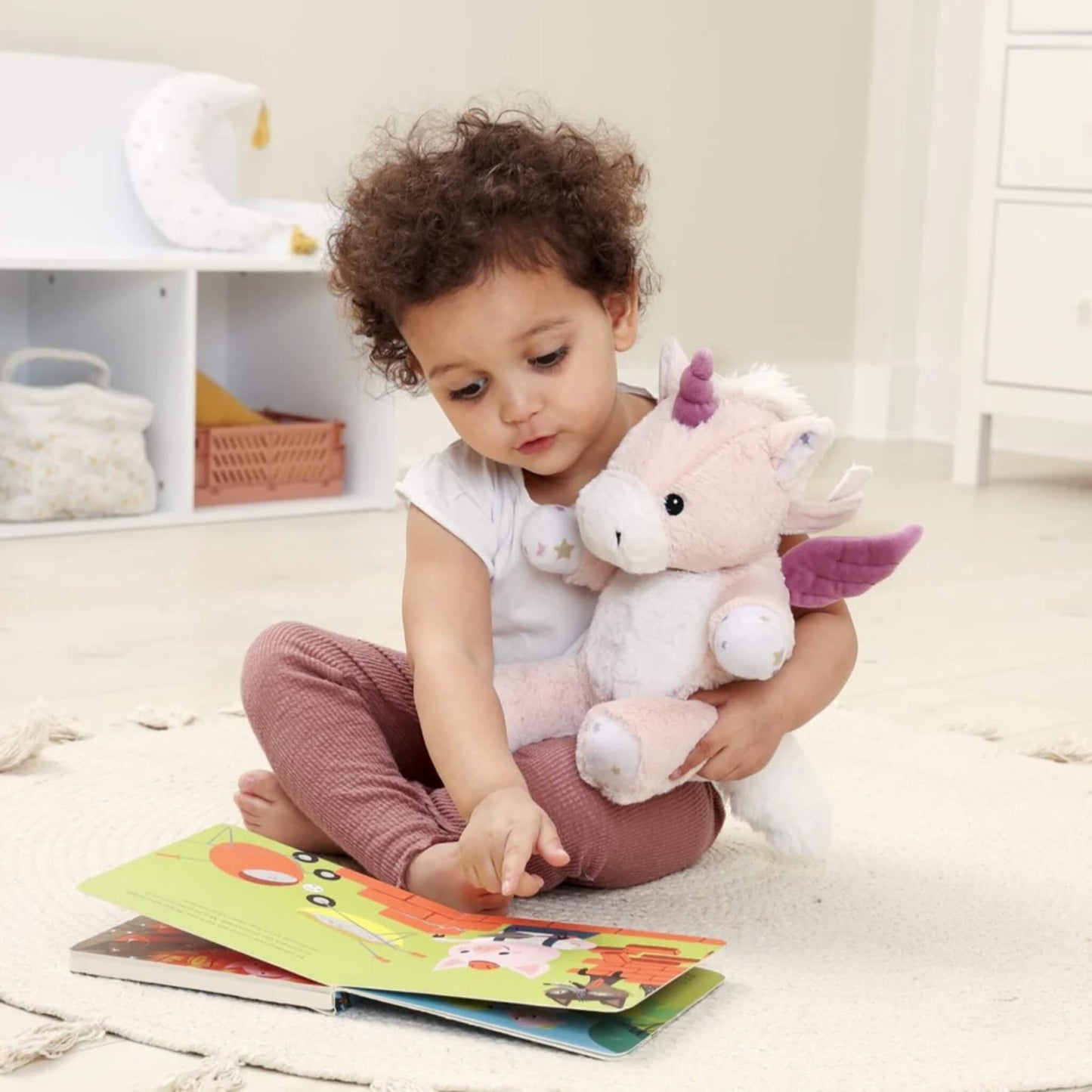 A young child with curly hair sits on a rug, pointing at a picture book while holding a plush unicorn toy with purple wings and a horn in their other arm.