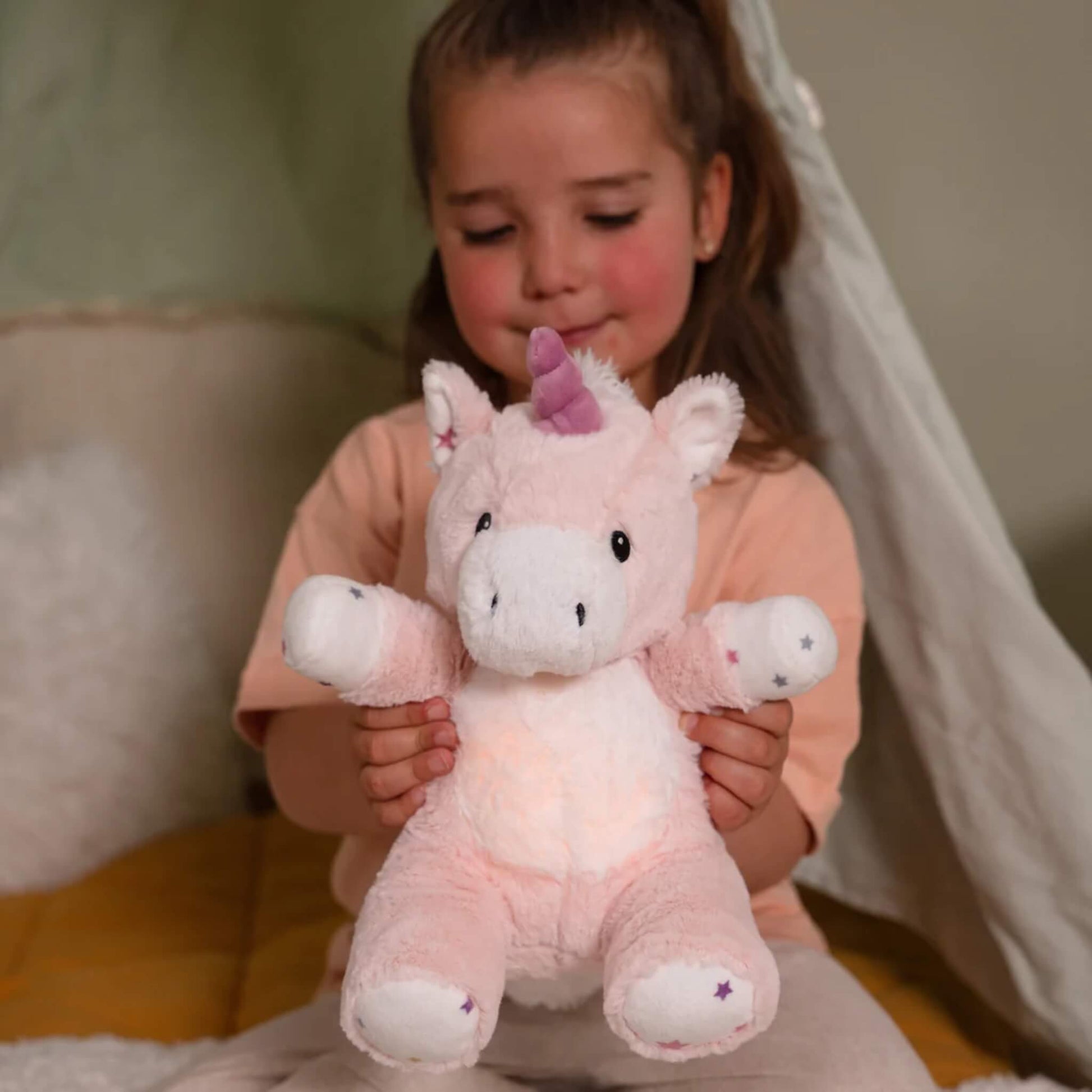 A girl with rosy cheeks holds a plush unicorn toy in her lap, the toy’s tummy softly glowing while she sits indoors against a draped background.