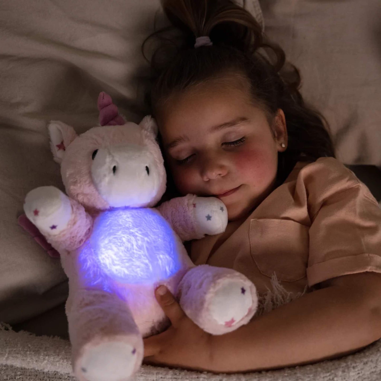 Child lying in bed hugging the glowing unicorn plush toy, with the tummy light shining blue in the dark.