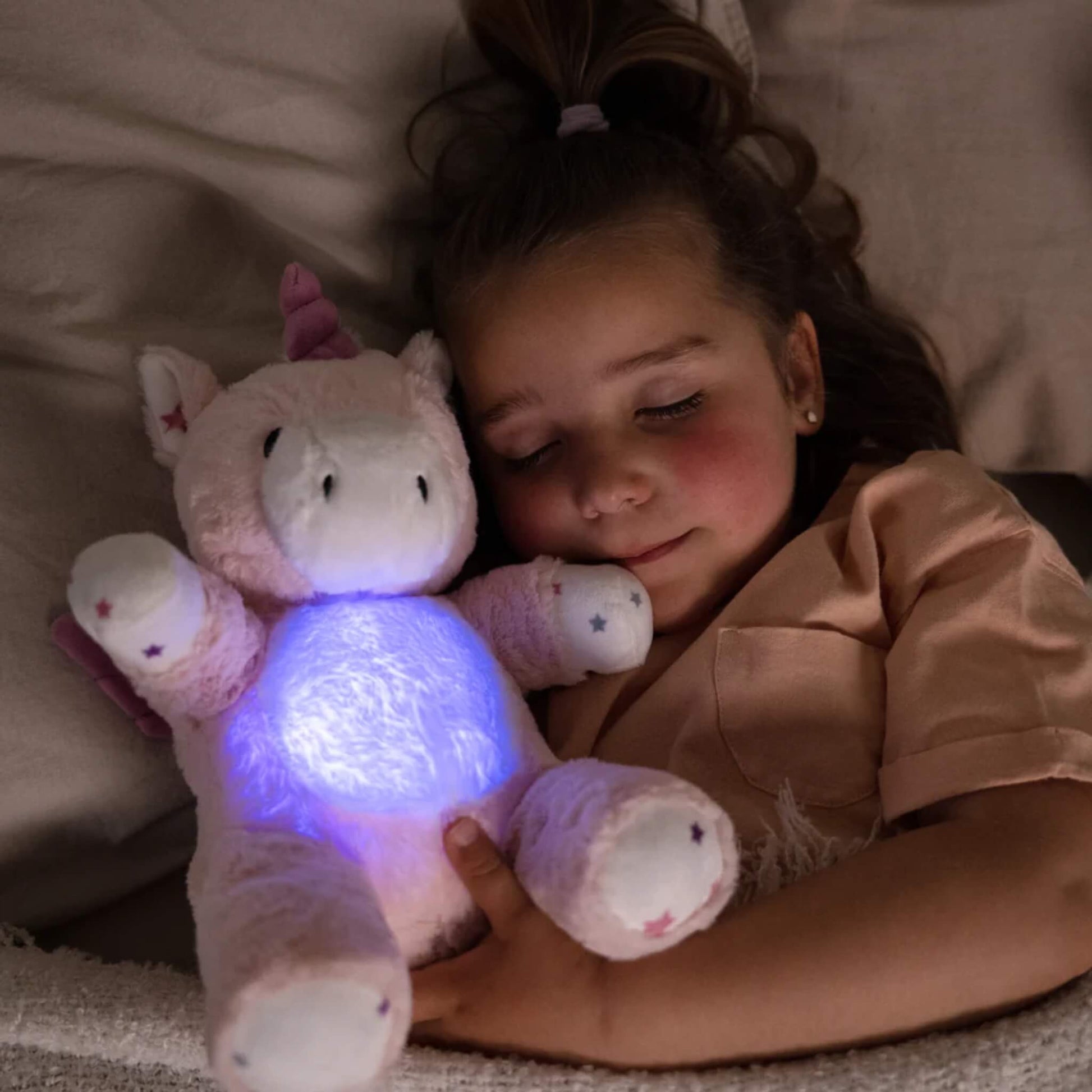 Child lying in bed hugging the glowing unicorn plush toy, with the tummy light shining blue in the dark.