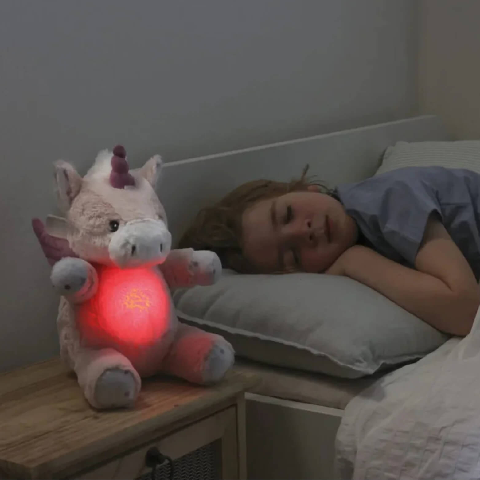 Unicorn plush toy placed on a bedside table, glowing red while a child sleeps next to it.