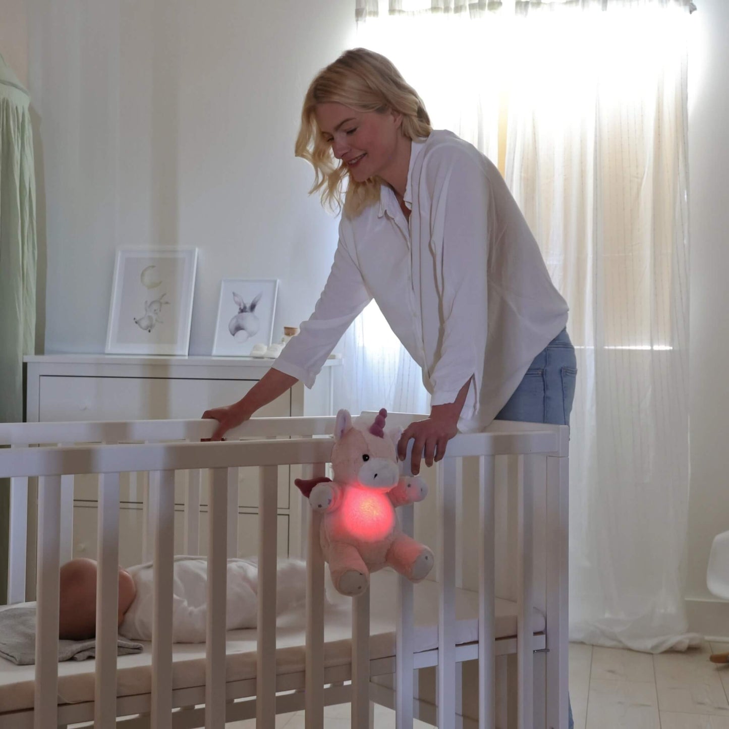Mother placing the unicorn plush toy on the side of a cot where a baby sleeps inside.