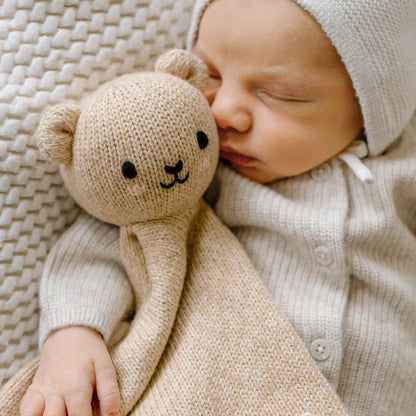 Sleeping newborn baby cuddled against a beige knitted bear comforter, showing the bear's embroidered face and soft organic cotton texture.