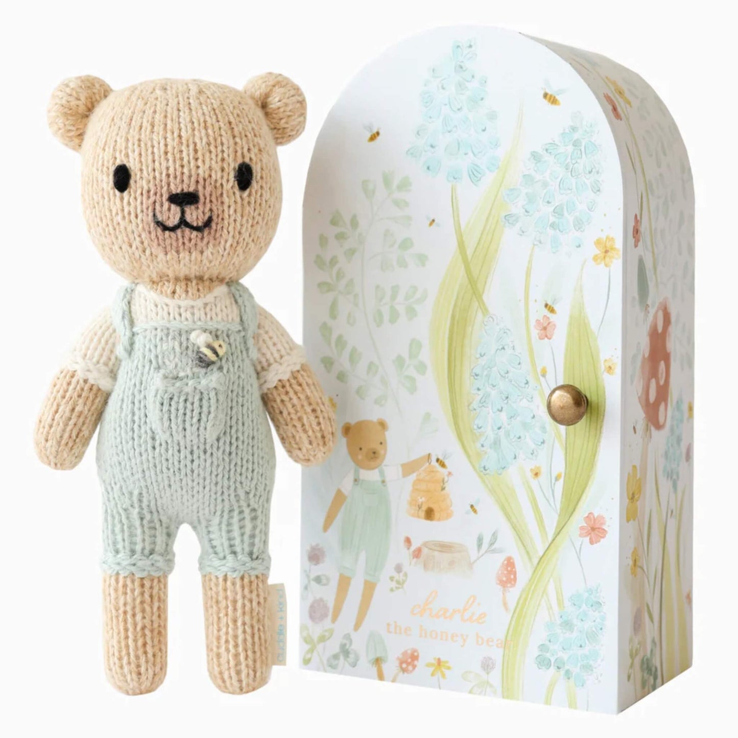 Cuddle + Kind Tiny Collection hand-knit doll in soft cotton with pale green overalls standing beside illustrated keepsake Tiny home box with floral design.
