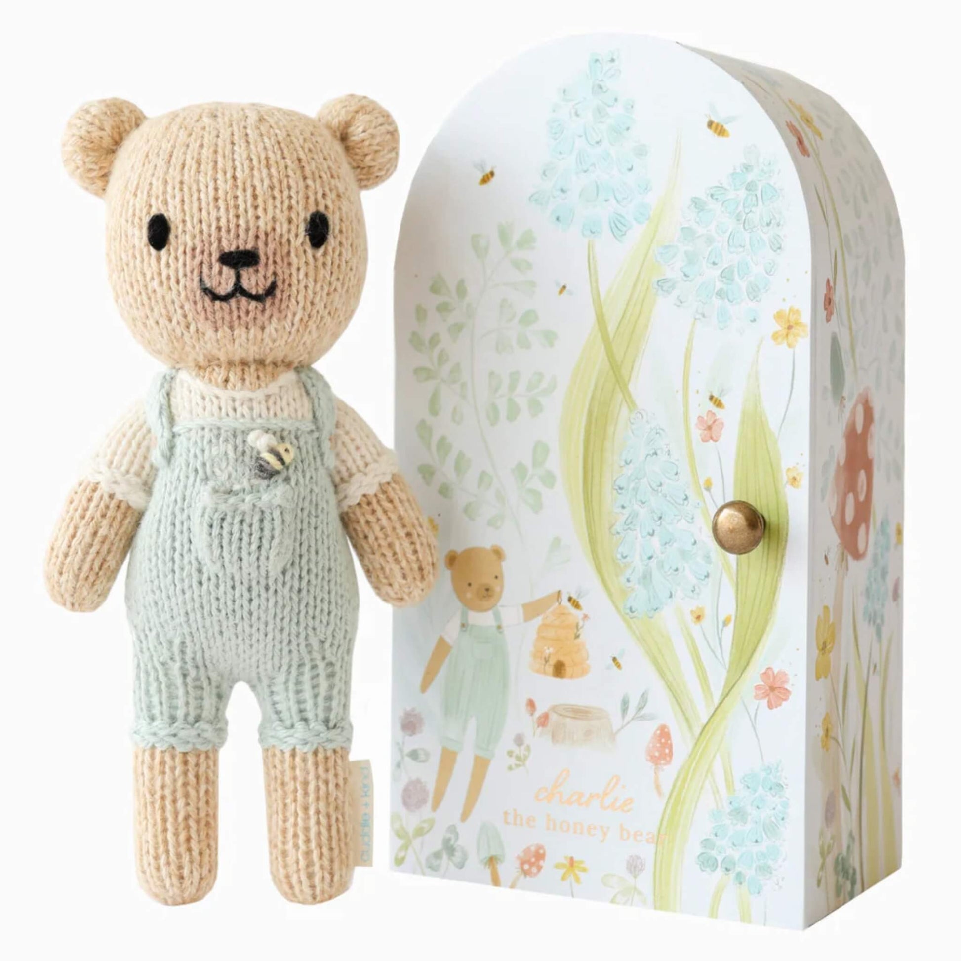 Cuddle + Kind Tiny Collection hand-knit doll in soft cotton with pale green overalls standing beside illustrated keepsake Tiny home box with floral design.