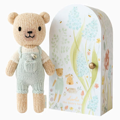 Cuddle + Kind Tiny Collection hand-knit doll in soft cotton with pale green overalls standing beside illustrated keepsake Tiny home box with floral design.