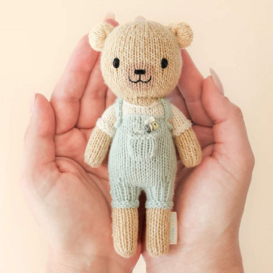 A small hand-knit cotton doll held gently in two hands, showing scale and soft stitched texture with pale green outfit.