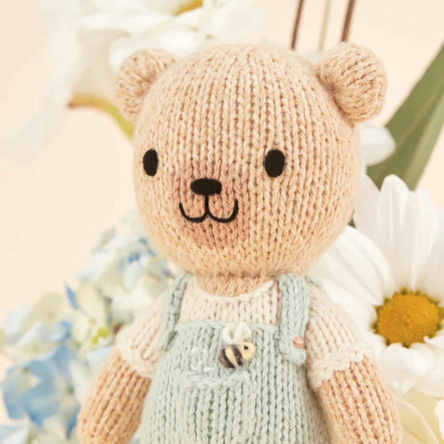 Close-up of a knitted doll’s face with soft stitching detail, neutral tones and gentle expression against a floral background.