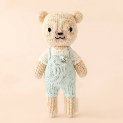 A hand-knit cotton doll standing upright against a neutral background, wearing pale green overalls with a small stitched detail on the chest.