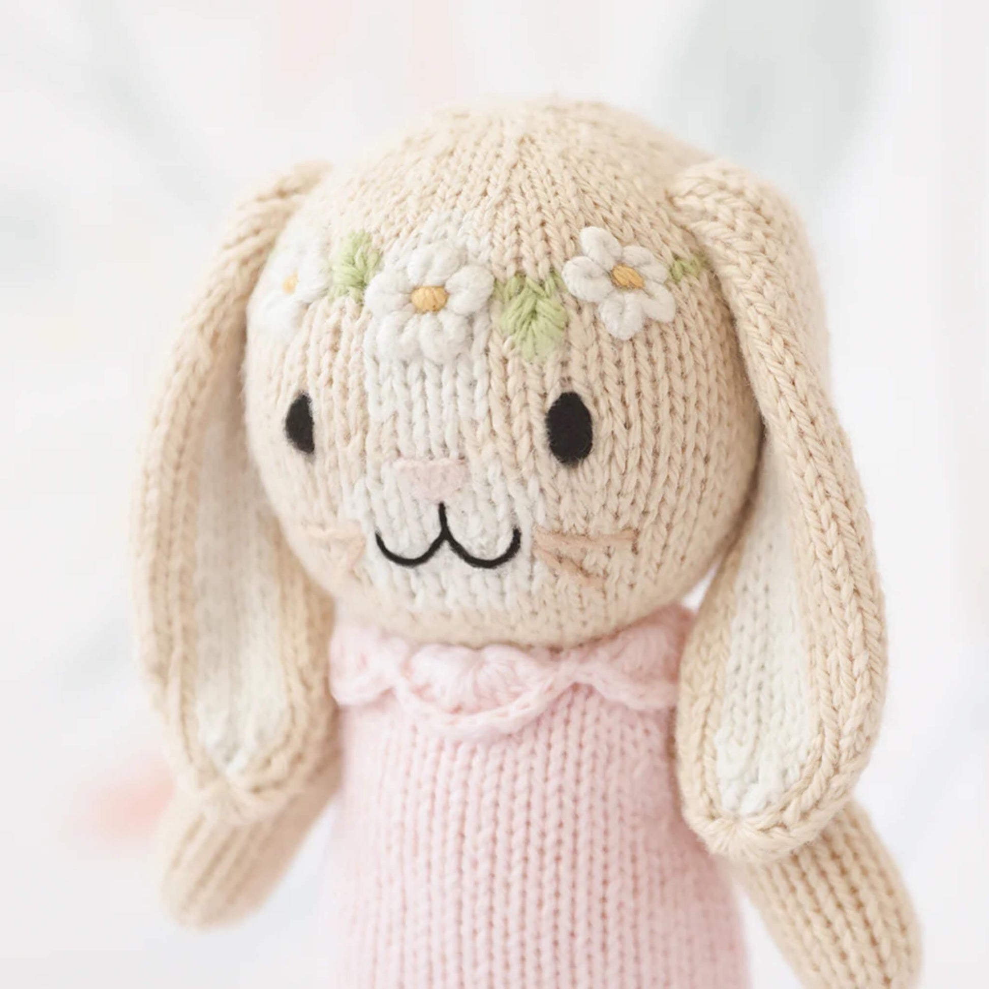 Close-up of knitted bunny face with stitched eyes, soft cream yarn texture and delicate flower crown detail.