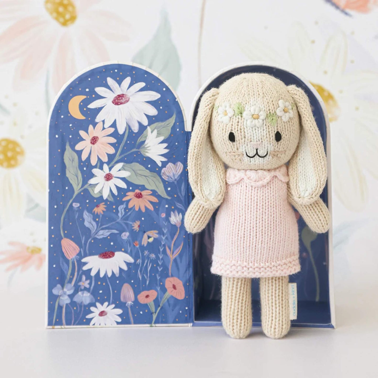 Knitted bunny doll displayed inside open gift box with illustrated night sky and floral interior design.