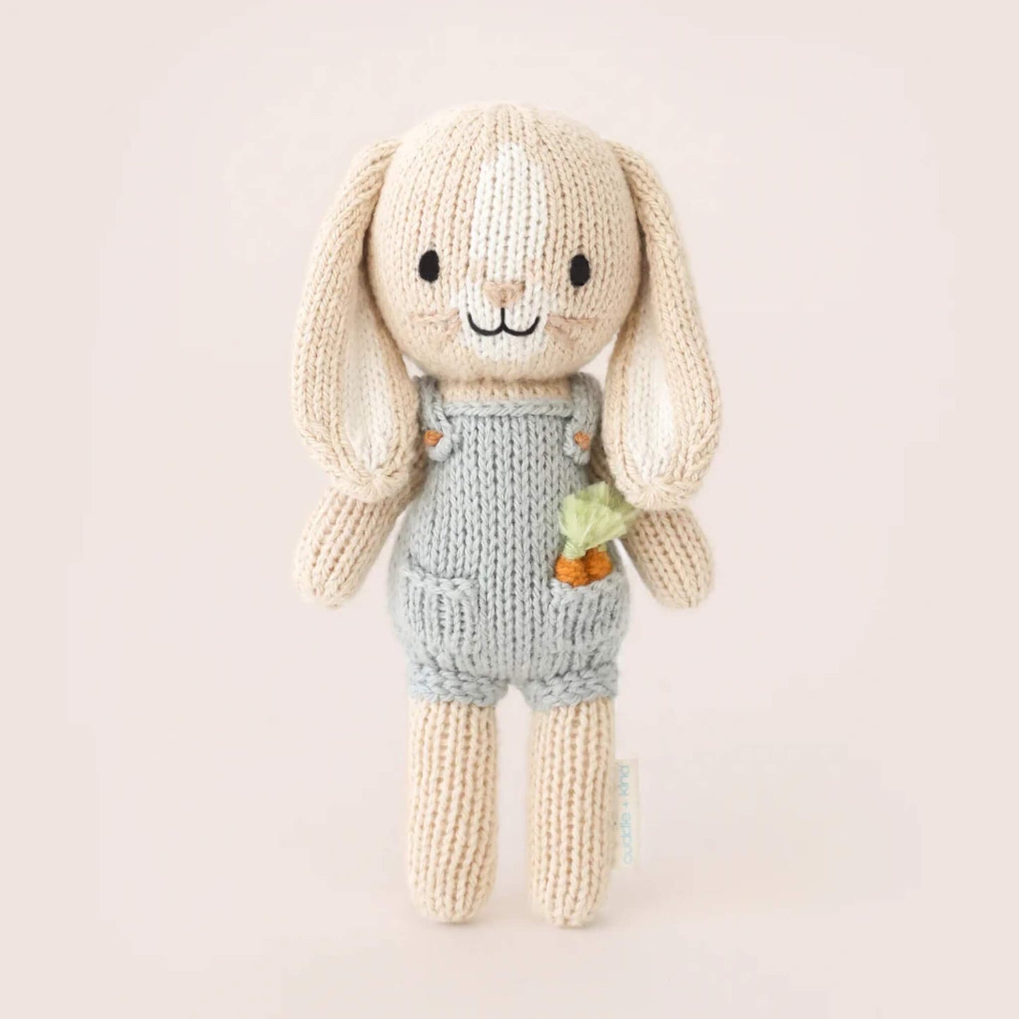 Full view of small knitted bunny soft toy standing upright, wearing grey overalls with textured knit finish and carrot detail.