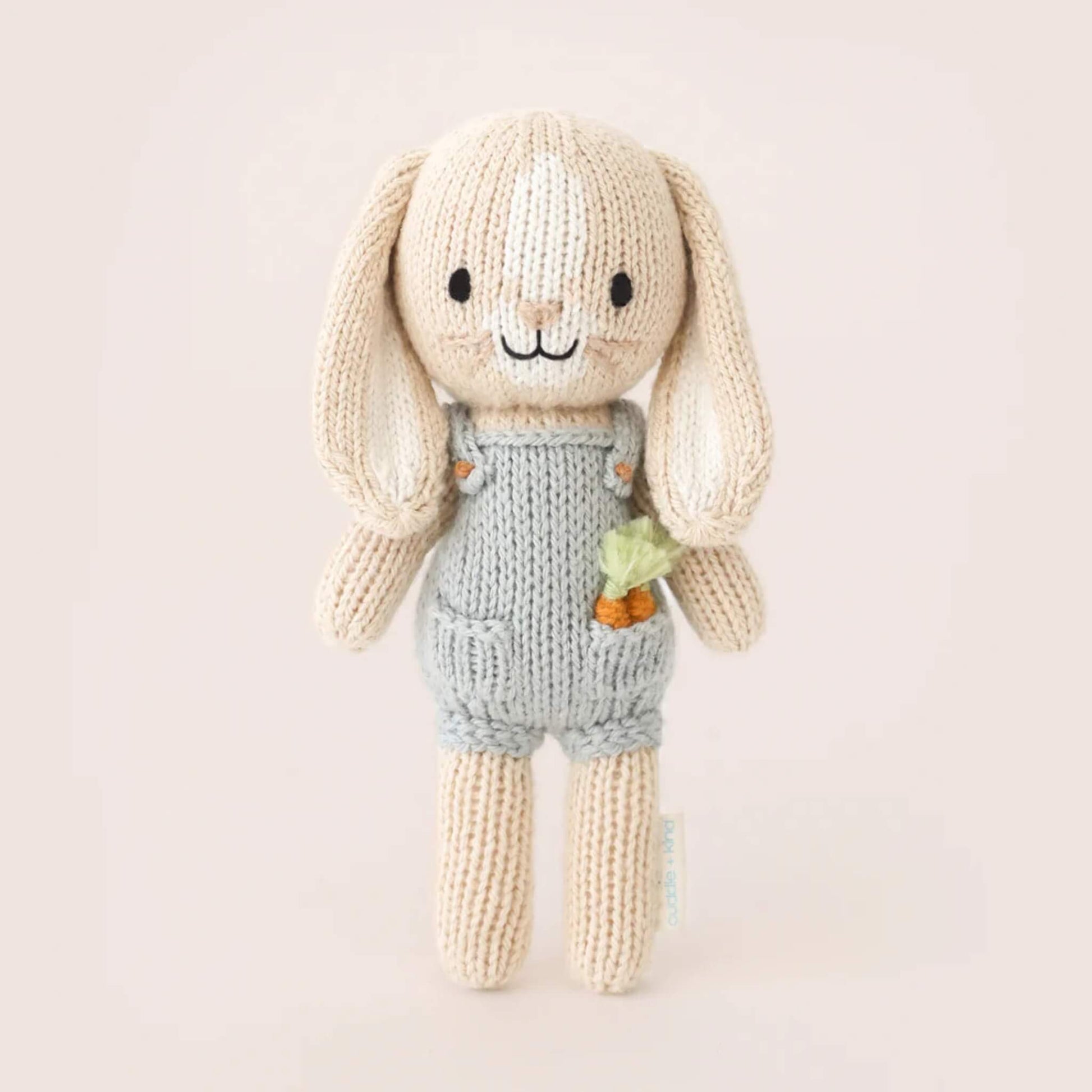 Full view of small knitted bunny soft toy standing upright, wearing grey overalls with textured knit finish and carrot detail.