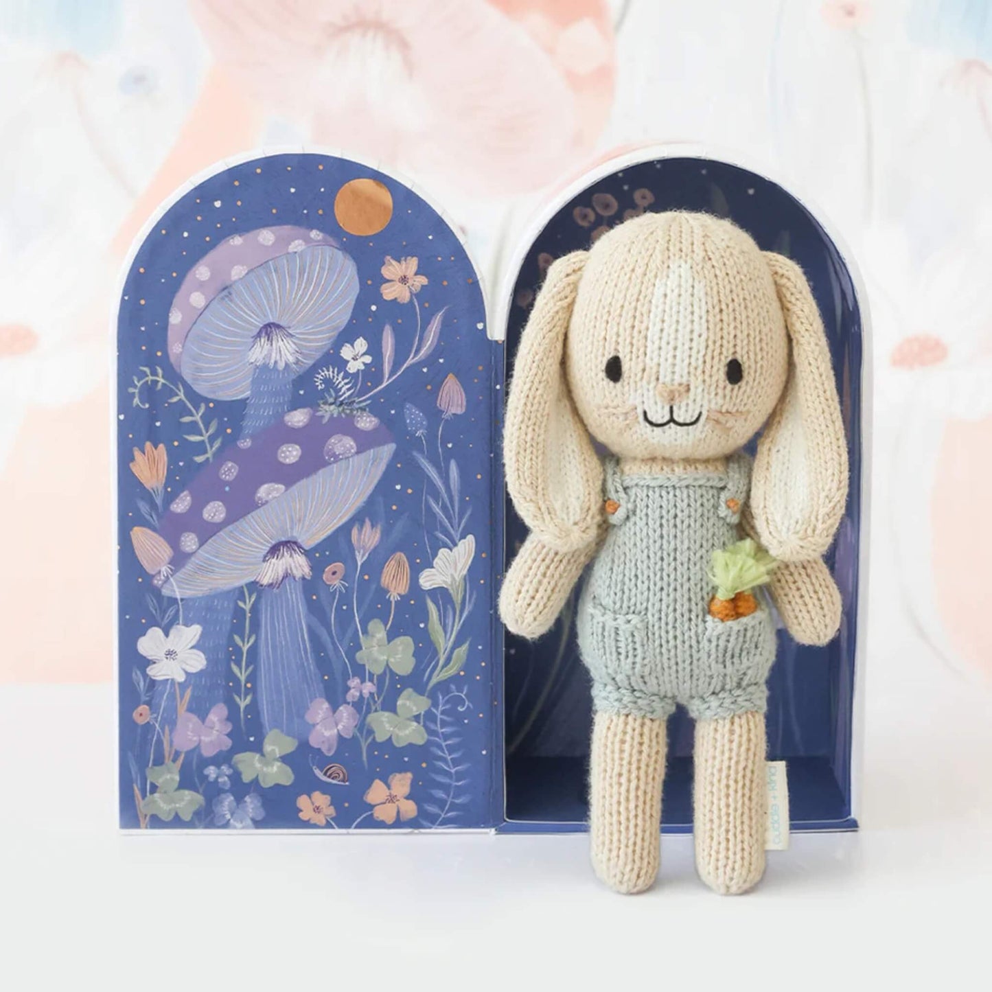 Knitted bunny displayed inside open gift box with dark blue illustrated interior showing night sky and mushrooms.