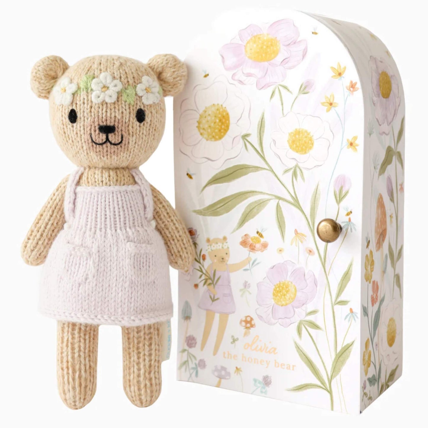Cuddle + Kind Tiny Collection hand-knit doll in soft cotton with pale pink dress and floral headband standing beside illustrated keepsake Tiny home box with botanical design.