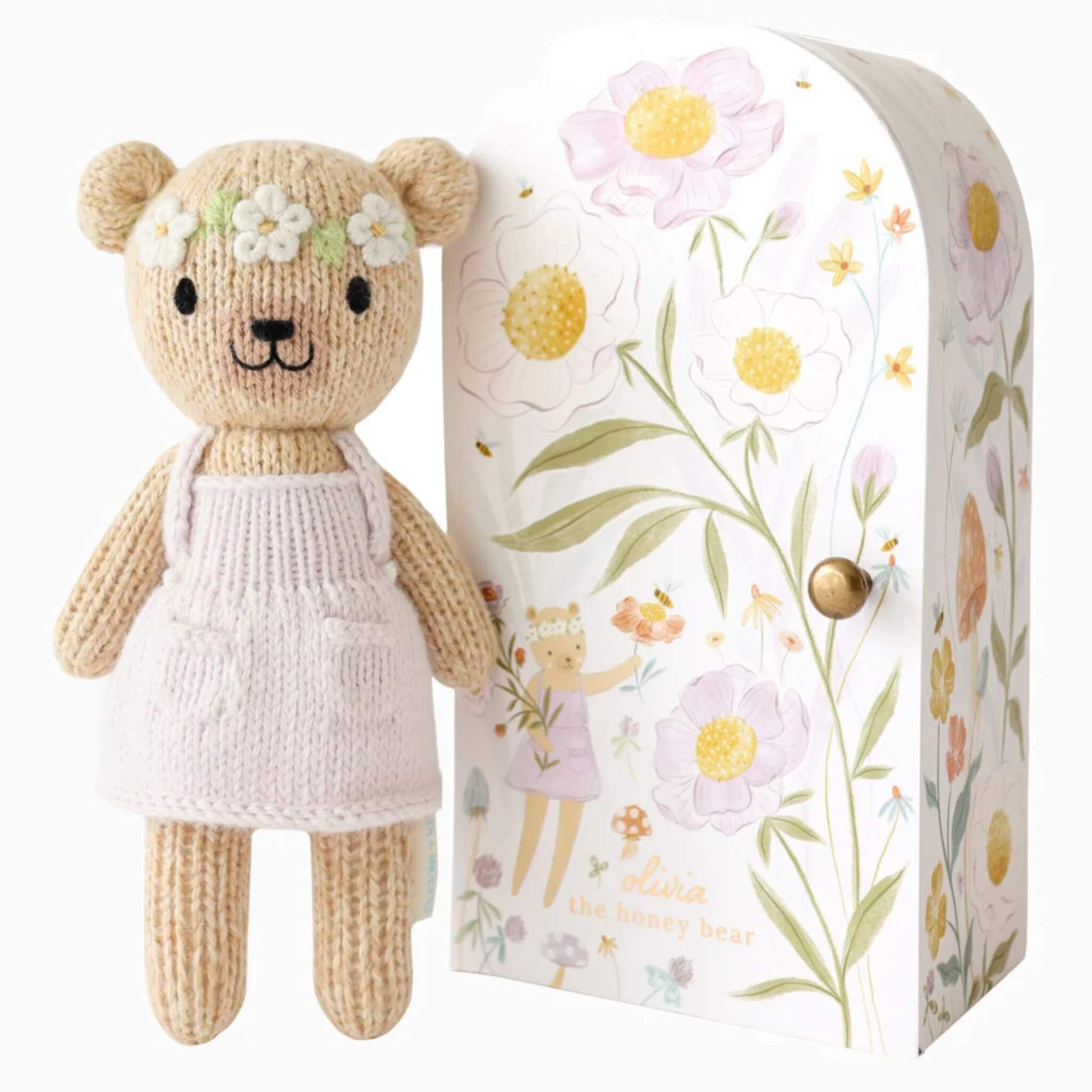 Cuddle + Kind Tiny Collection hand-knit doll in soft cotton with pale pink dress and floral headband standing beside illustrated keepsake Tiny home box with botanical design.