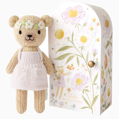 Cuddle + Kind Tiny Collection hand-knit doll in soft cotton with pale pink dress and floral headband standing beside illustrated keepsake Tiny home box with botanical design.