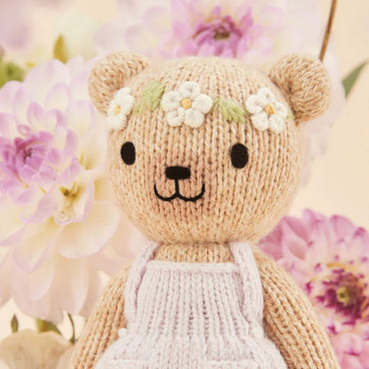Close-up of a knitted doll’s face with embroidered features and floral headband, set against a soft floral background.