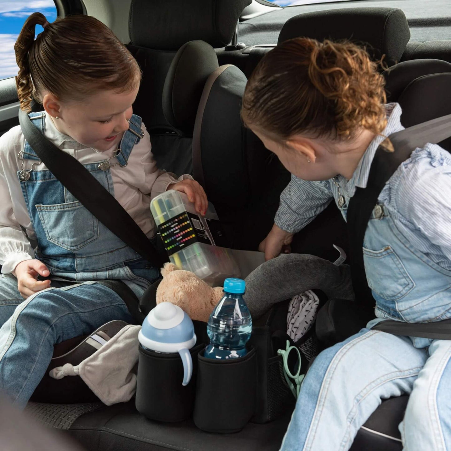 Two children in car seats looking into a black car seat basket positioned between them, filled with drinks, toys, and activity supplies.