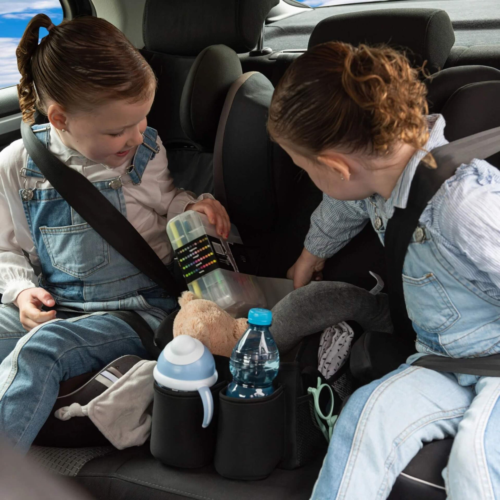 Two children in car seats looking into a black car seat basket positioned between them, filled with drinks, toys, and activity supplies.