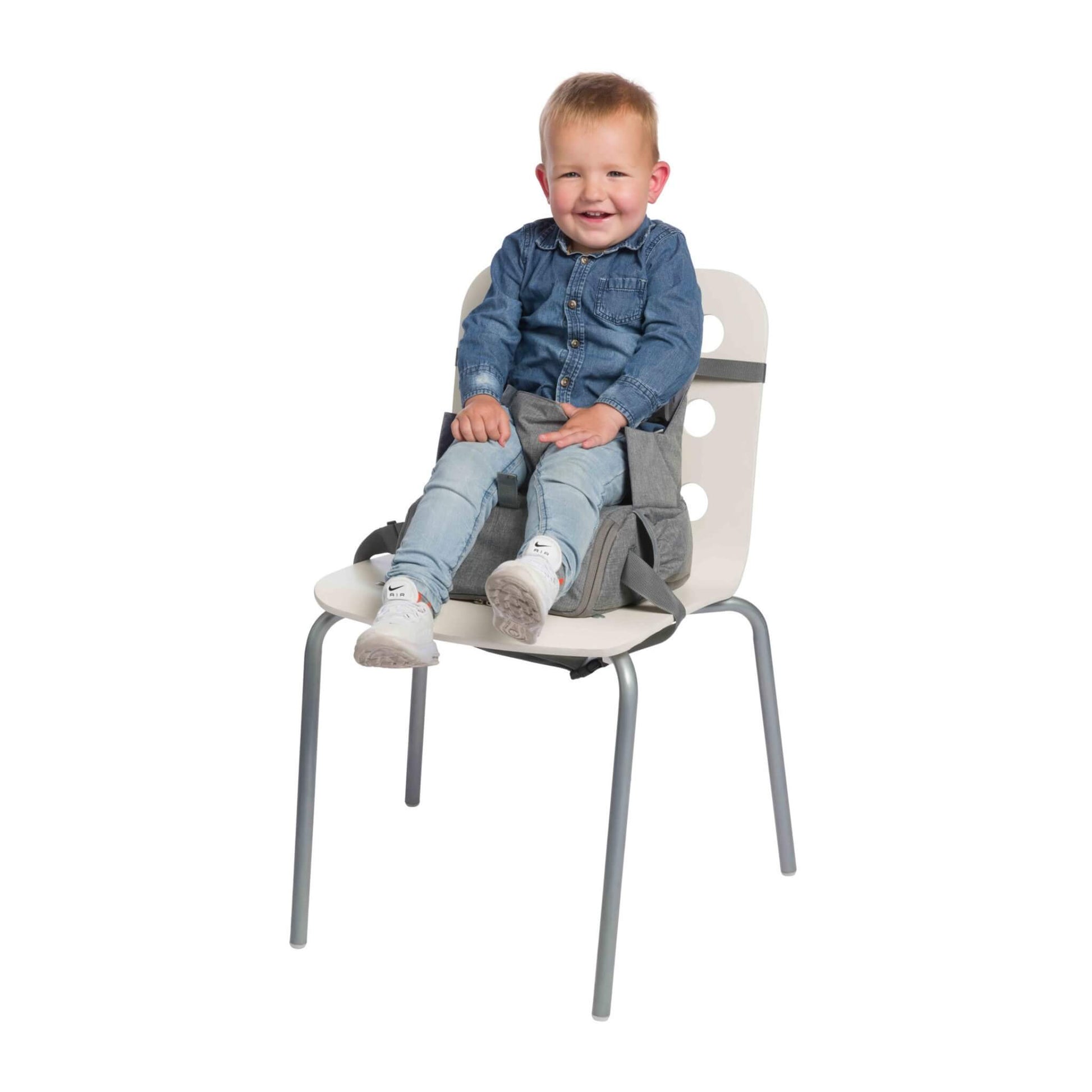 A toddler sitting in a grey booster seat secured to a white chair with a safety harness.