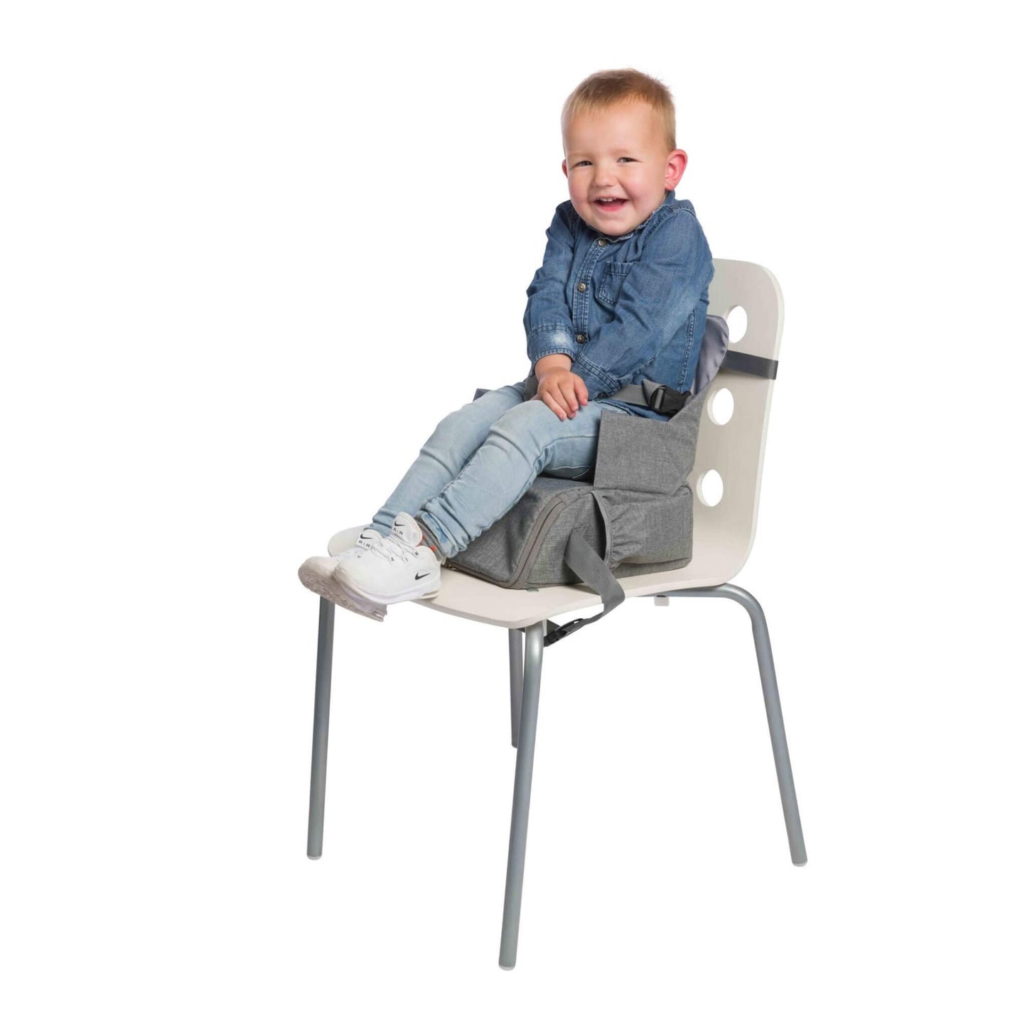 A toddler smiling while sitting on a grey booster seat placed on a white chair.
