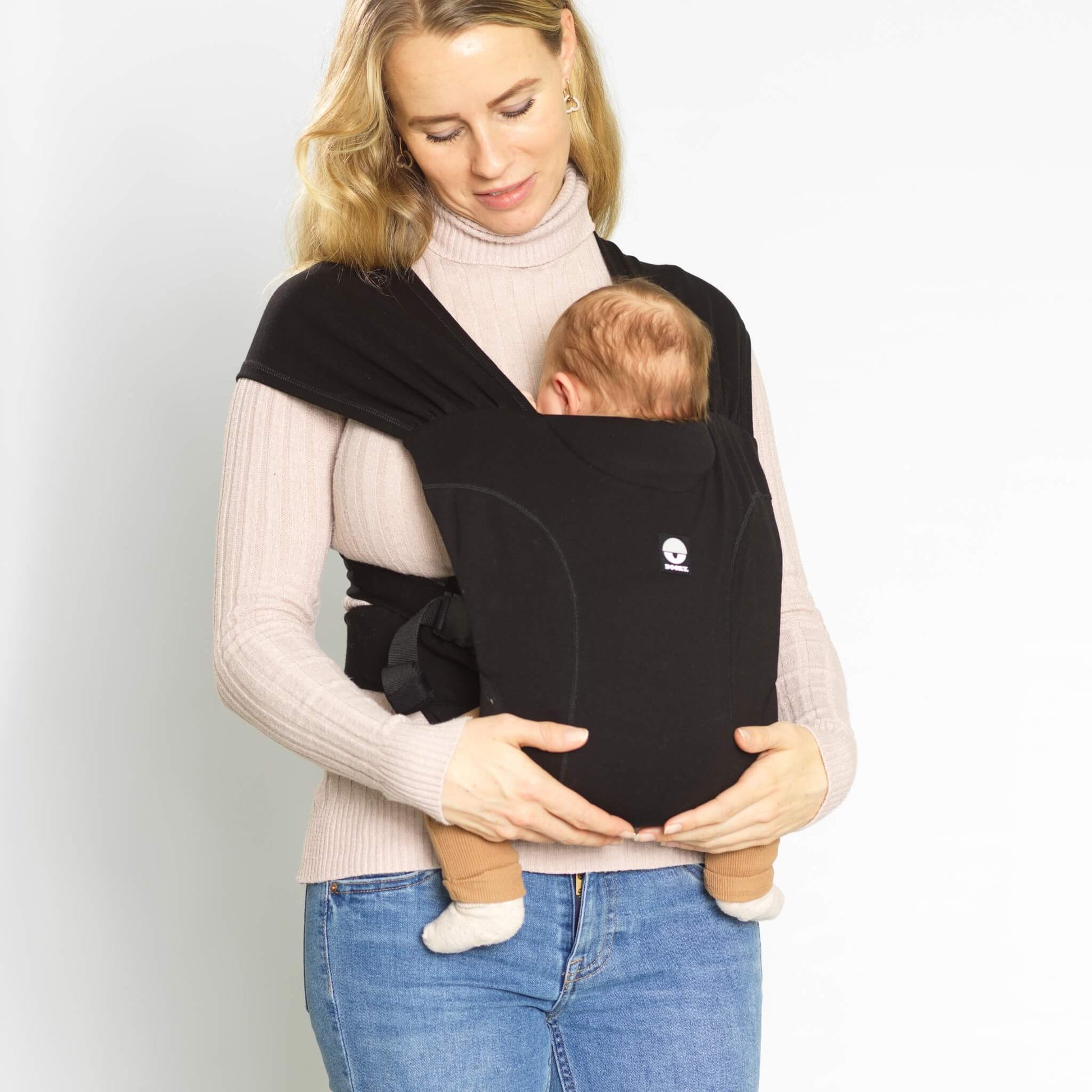 A woman wearing a black baby carrier with a young baby carried close against her chest in a supported position.