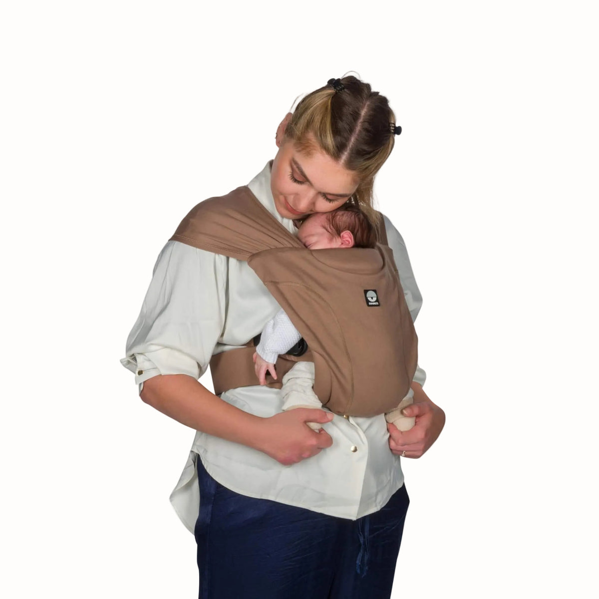 Woman wearing a mocha baby carrier with a newborn resting upright against her chest and head nestled beneath the shoulder strap.