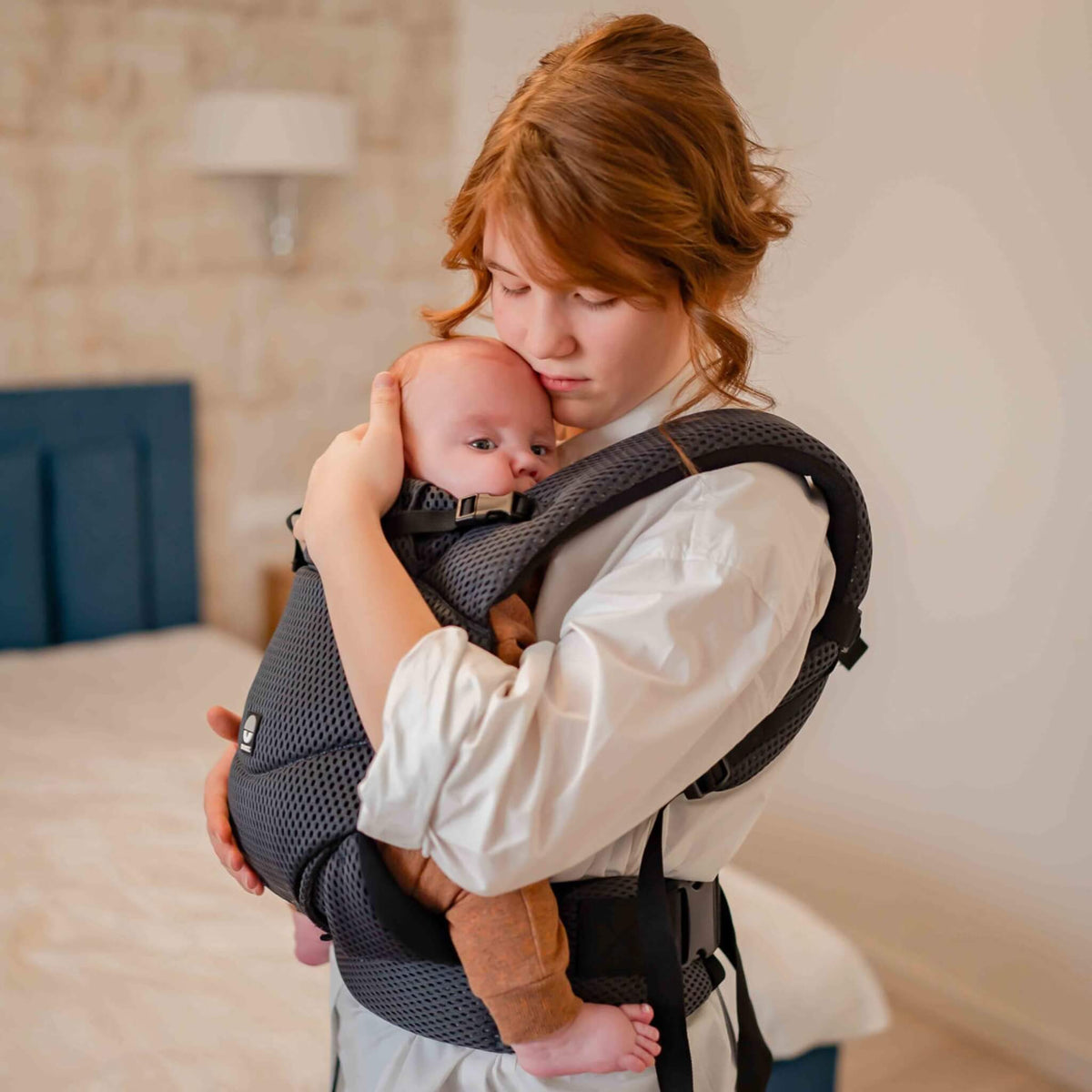 A woman carrying a young baby close to her chest in a grey breathable mesh baby carrier, in a bedroom setting.