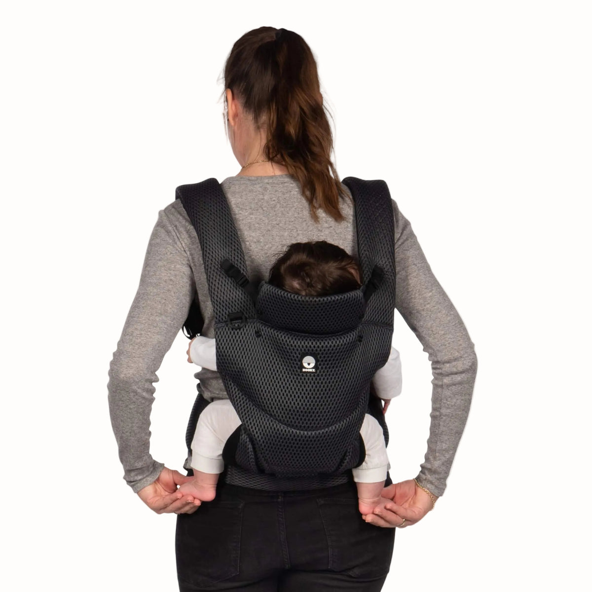 Rear view of a woman carrying a baby in a grey mesh baby carrier, showing the padded shoulder straps and supportive back panel.