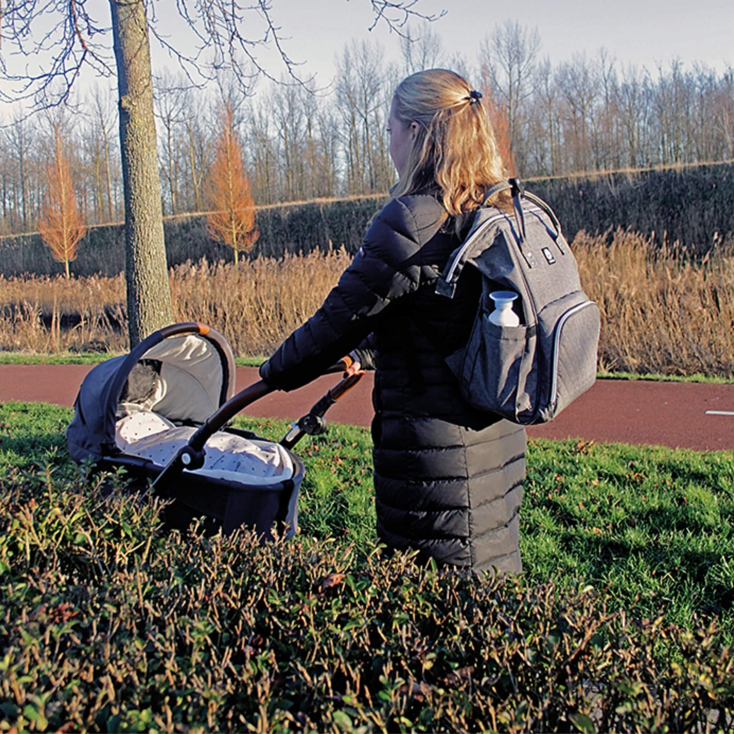 A person wearing a grey changing bag as a backpack while pushing a pram along an outdoor path on a winter day, with a bottle visible in the side pocket.