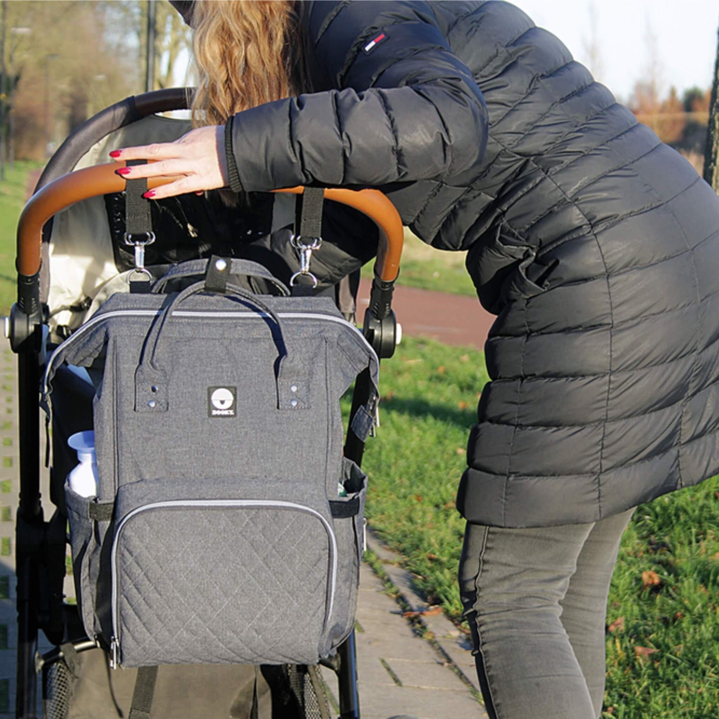 A person leaning over a pushchair with a grey changing bag securely clipped to the handlebar using two black stroller straps.