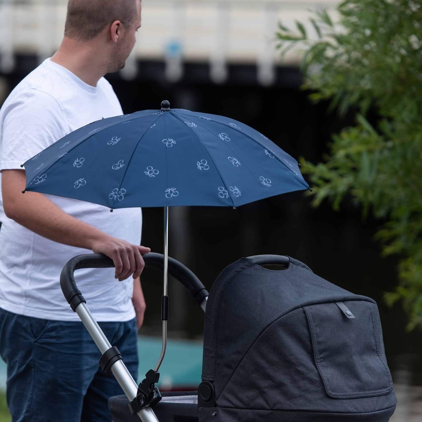 Parent walking outdoors with the Dooky Blue Cherry parasol attached to a pram, offering sun protection on a sunny day.