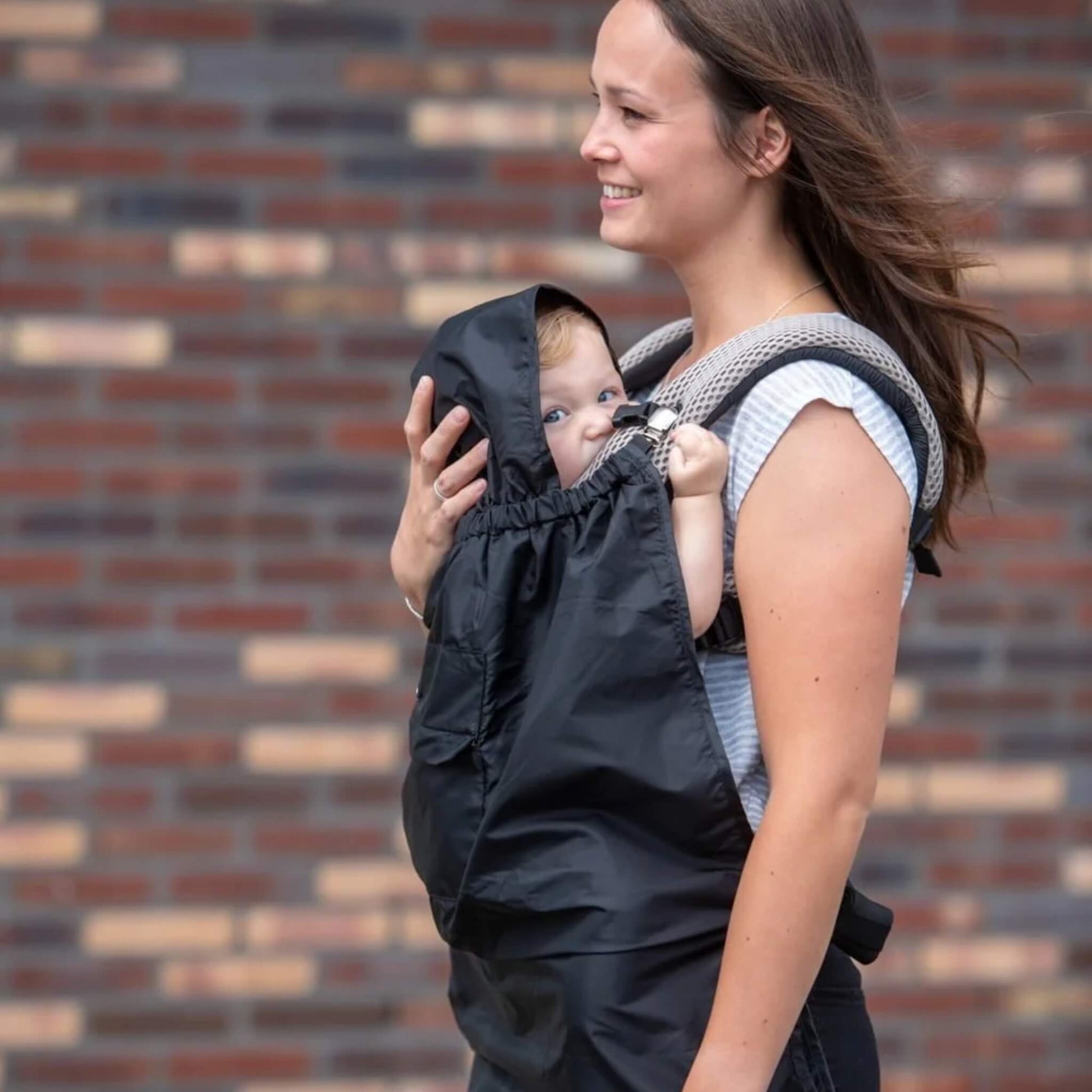 Close-up side view of a woman walking outdoors wearing a baby in a carrier covered by a black rain cape, with the baby looking out from under the hood.
