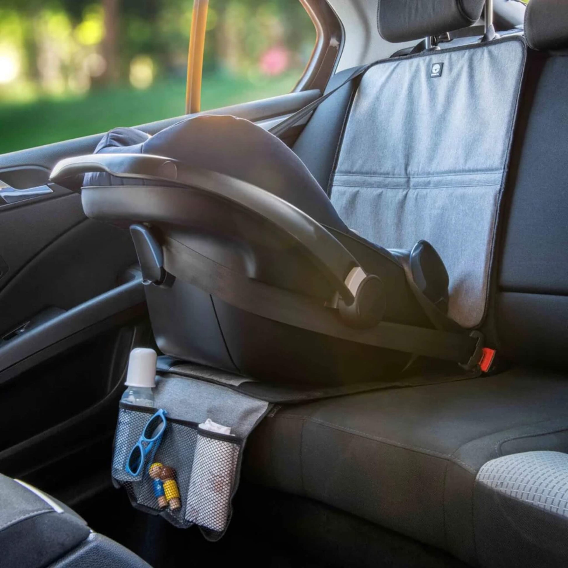 A grey and black seat protection mat installed in a car with an infant car seat secured on top and small items stored in the mesh pockets.