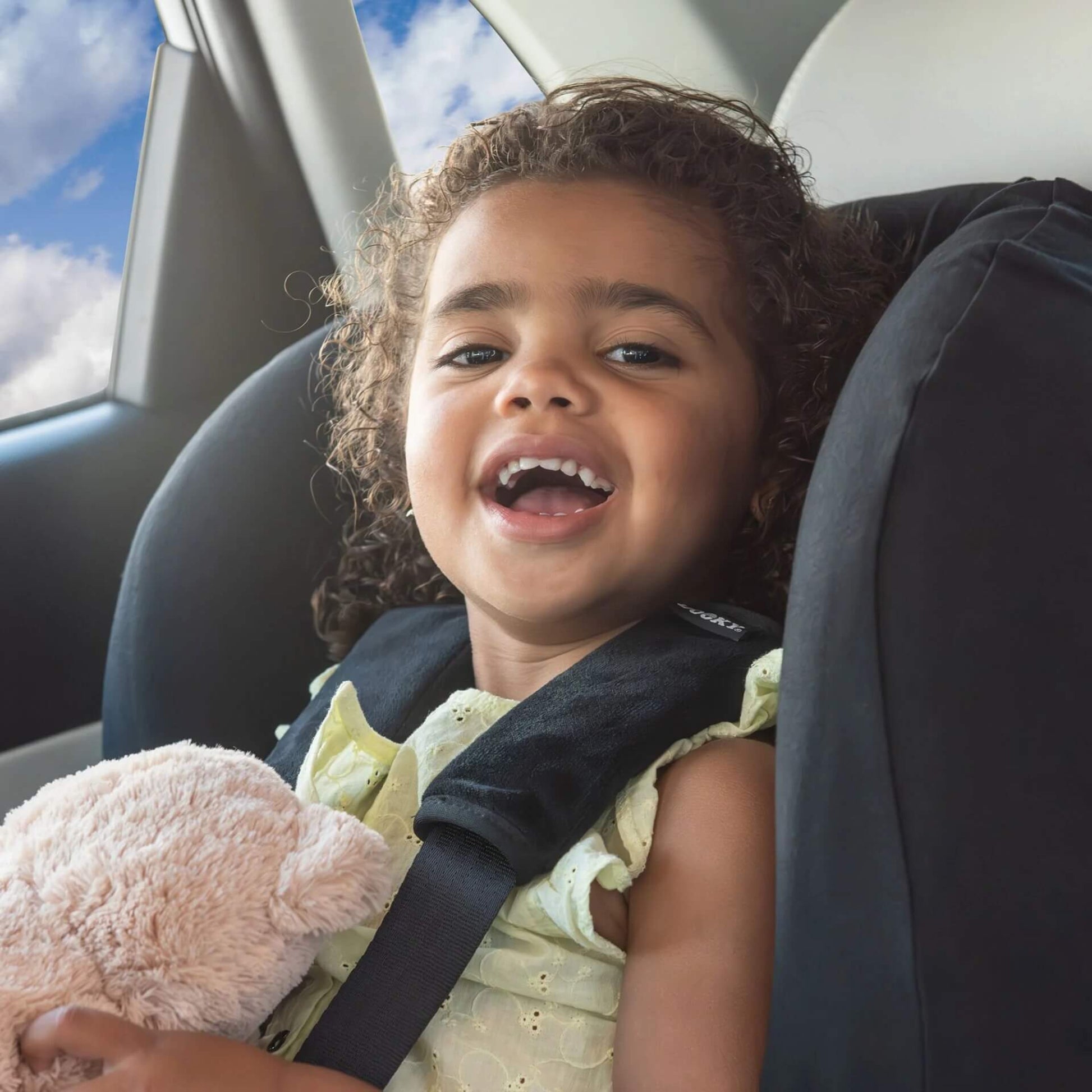 A young child sitting in a car seat wearing a light yellow dress, smiling and holding a soft toy, with black padded seatbelt covers fitted to the harness straps.